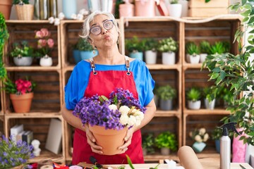 Middle age woman with grey hair working at florist shop holding plant looking at the camera blowing a kiss being lovely and sexy. love expression.