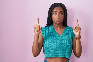 Young african american with braids standing over pink background pointing up looking sad and upset, indicating direction with fingers, unhappy and depressed.