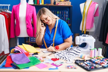 Fototapeta premium Young woman tailor talking on smartphone writing on notebook at sewing studio