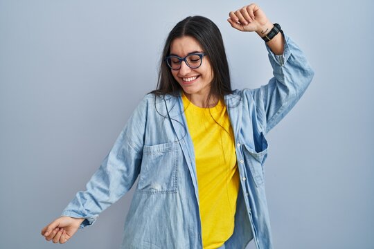 Young hispanic woman standing over blue background dancing happy and cheerful, smiling moving casual and confident listening to music
