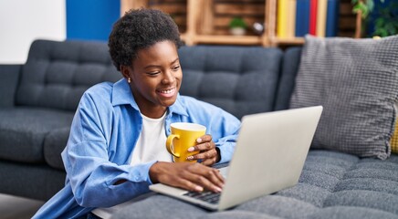 African american woman using laptop and drinking coffee sitting on floor at home