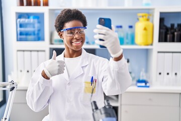 African american woman working at scientist laboratory doing video call with smartphone smiling happy and positive, thumb up doing excellent and approval sign