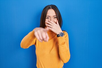 Young brunette woman standing over blue background laughing at you, pointing finger to the camera...