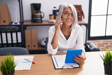 Middle age grey-haired woman business worker talking on smartphone using touchpad at office