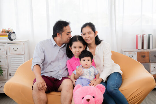 Selective focus Asian mother ans father sitting on cozy sofa cuddling and kissing with two little children take a photo in living room. Sibling with mom and dad spend time together in holiday
