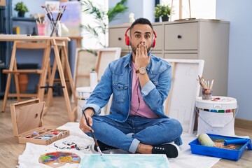Young hispanic man painter sitting on the floor at art studio covering mouth with hand, shocked and...