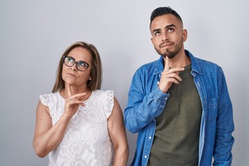 Hispanic mother and son standing together thinking concentrated about doubt with finger on chin and looking up wondering