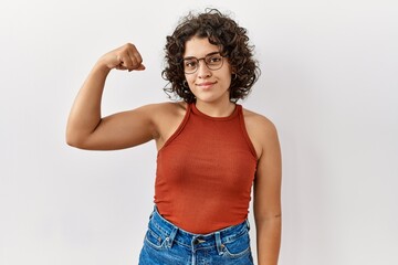 Young hispanic woman wearing glasses standing over isolated background strong person showing arm muscle, confident and proud of power