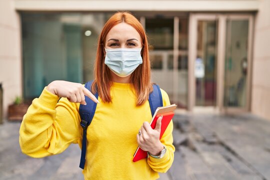 Young woman wearing safety mask and student backpack holding books pointing finger to one self smiling happy and proud