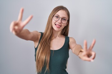 Young caucasian woman standing over white background smiling with tongue out showing fingers of both hands doing victory sign. number two.