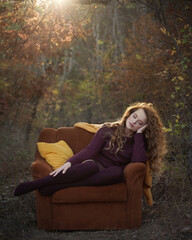 A beautiful young red -haired woman sitting in a cozy armchair with pillows against the background of an autumn misty forest in the sunset light .