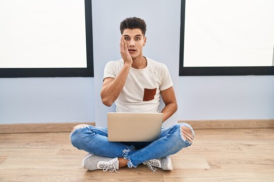 Young hispanic man using laptop at home afraid and shocked, surprise and amazed expression with hands on face
