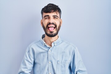 Young hispanic man with beard standing over blue background sticking tongue out happy with funny expression. emotion concept.