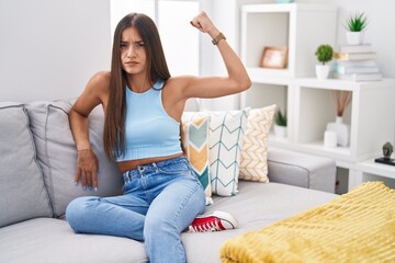 Young brunette woman sitting on the sofa at home strong person showing arm muscle, confident and proud of power