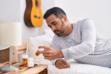 Young hispanic man lying on bed holding pills at bedroom