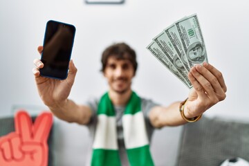 Naklejka premium Young hispanic man holding smartphone and dollars at home