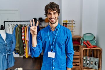 Young hispanic man working as manager at retail boutique showing and pointing up with fingers number three while smiling confident and happy.