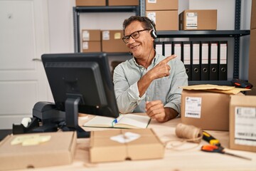 Senior man working at small business ecommerce wearing headset smiling cheerful pointing with hand and finger up to the side