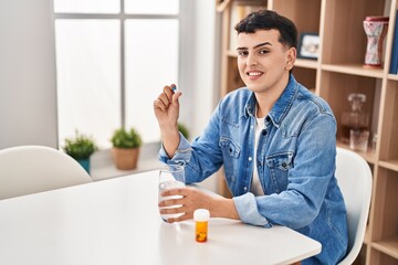 Young non binary man taking pill sitting on table at home