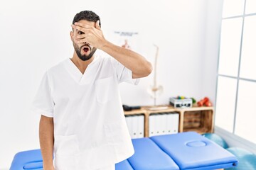 Young handsome man with beard working at pain recovery clinic peeking in shock covering face and eyes with hand, looking through fingers with embarrassed expression.