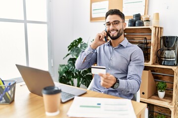 Young arab man talking on the smartphone holding credit card working at office