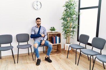 Young arab man sitting on chair with arm sling at clinic waiting room