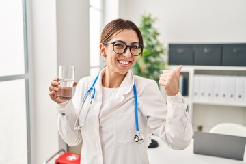 Young brunette doctor woman holding glass of water smiling happy pointing with hand and finger to the side