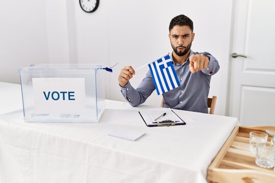 Young handsome man with beard at political campaign election holding greece flag pointing with finger to the camera and to you, confident gesture looking serious