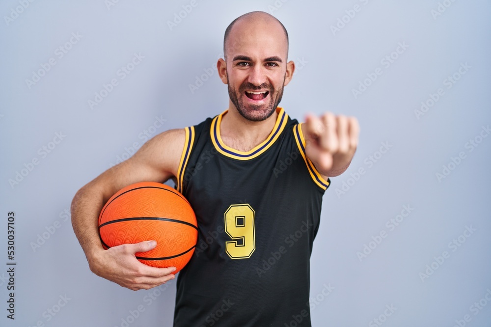 Poster young bald man with beard wearing basketball uniform holding ball pointing to you and the camera wit