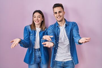 Young hispanic couple standing over pink background smiling cheerful offering hands giving assistance and acceptance.