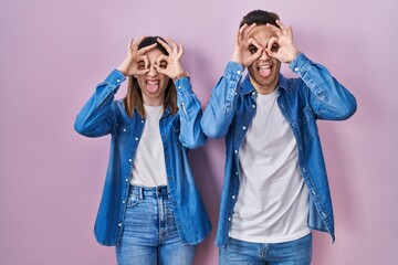 Fototapeta na wymiar Young hispanic couple standing over pink background doing ok gesture like binoculars sticking tongue out, eyes looking through fingers. crazy expression.