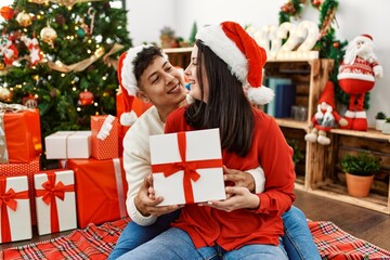Obraz na płótnie Canvas Young hispanic couple smiling happy and hugging. Sitting on the floor wearing christmas hat holding gift at home.