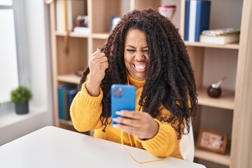 Plus size hispanic woman using smartphone sitting on the table screaming proud, celebrating victory...