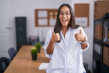 Young hispanic woman at the office success sign doing positive gesture with hand, thumbs up smiling...