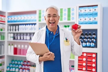 Poster Middle age man with grey hair working at pharmacy drugstore holding red heart smiling and laughing hard out loud because funny crazy joke. © Krakenimages.com