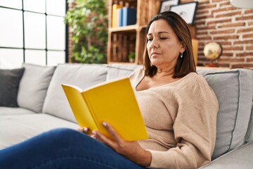 Middle age hispanic woman reading book sitting on sofa at home