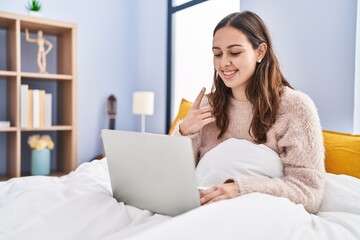 Young hispanic woman using computer laptop on the bed smiling happy pointing with hand and finger