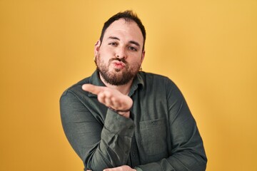 Plus size hispanic man with beard standing over yellow background looking at the camera blowing a kiss with hand on air being lovely and sexy. love expression.