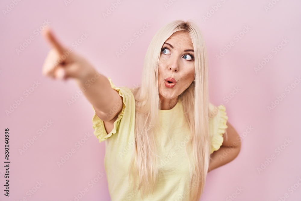 Poster caucasian woman standing over pink background pointing with finger surprised ahead, open mouth amaze