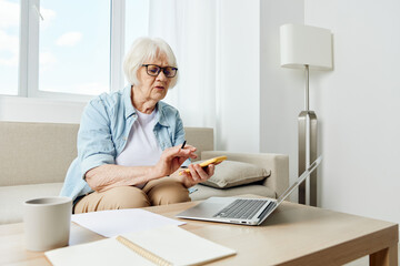 a beautiful elderly woman who is passionate about work is sitting at home in a bright apartment on a cozy sofa and checking finances looking at a laptop doing calculations on a calculator in her phone