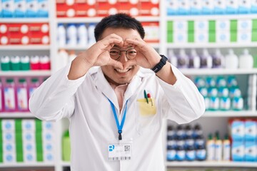 Chinese young man working at pharmacy drugstore doing heart shape with hand and fingers smiling looking through sign