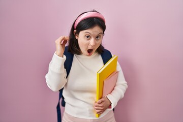 Woman with down syndrome wearing student backpack and holding books in shock face, looking skeptical and sarcastic, surprised with open mouth