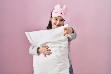 Woman with down syndrome wearing sleeping mask hugging pillow pointing to you and the camera with fingers, smiling positive and cheerful