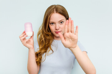 Young caucasian woman holding deodorant isolated on blue background standing with outstretched hand showing stop sign, preventing you. - obrazy, fototapety, plakaty