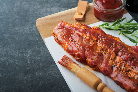 Pork ribs. Raw meat. Raw pork ribs in marinade with spices, rosemary, tomato sauce and garlic on white marble stand on black stone table background. Barbecue concept. Selective focus. Mock up.