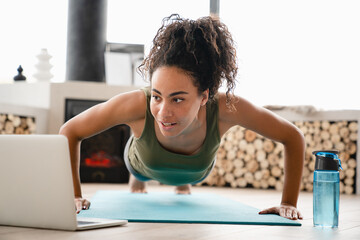 Fit healthy african young woman athlete doing yoga, training, workout push-ups on fitness mat watching online tutorial on laptop at home