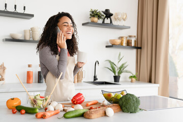 Obraz na płótnie Canvas African young woman housewife chef cooking dinner, making vegetable vegan salad at home kitchen while talking on cellphone with friends discussing recipe
