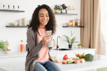 Cheerful african young woman housewife freelancer relaxing at home kitchen, reading news online, using cellphone for mobile application, social media
