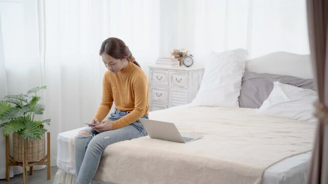 Asian woman sitting on bed using smartphone chatting with bestfriend,  young girl takes a break from working with laptop to play on mobile phone. free time after work.
