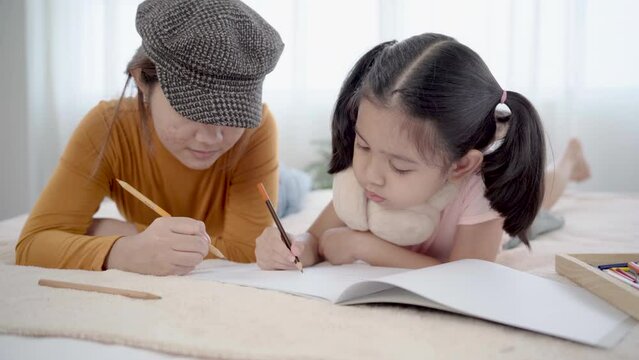 Sibling drawing book together on bed, elder sister and adorable little girl enjoying hobby pastime alone without parents in bedroom. Nanny teaching children draw in holiday, using colorful pencils
